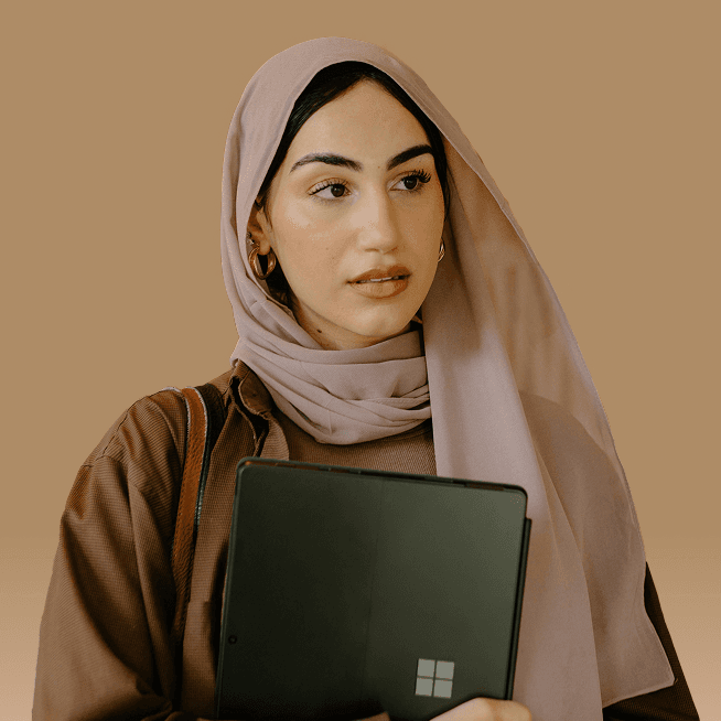 A person wearing a light pink hijab and brown top holding a tablet against a neutral beige background.