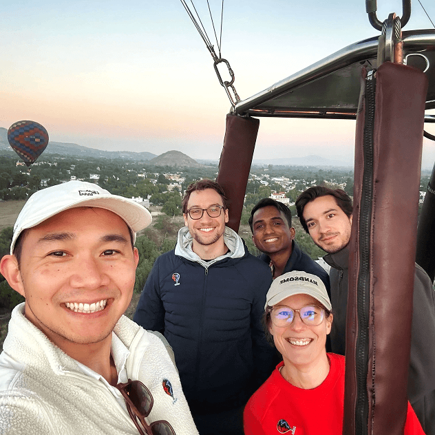 Five people taking a selfie from a hot air balloon basket with aerial views of a town and other hot air balloons visible in the distance.